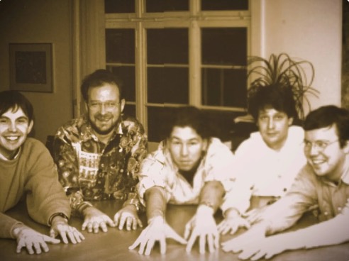 The founding team around a table in 1986, hands flat on the wood.
